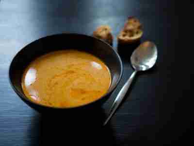 Homemade hot orange crab bisque in black ceramic bowl with slices of grilled bread and a spoon, placed on black wooden table
