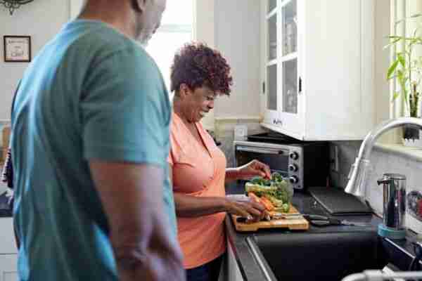 Man watching with focus on woman in casual clothing standing at kitchen counter chopping vegetables.