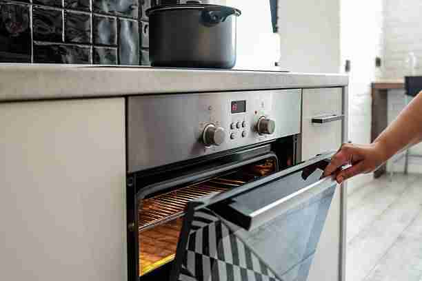 Close-up of a hand opening a hot oven in a modern kitchen. The oven is glowing inside, ready for baking. A black pot is seen on the stovetop above.