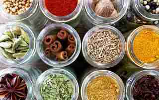 A selection of dried spices and herbs in glass jars shot on a dark wood background. Coriander seed, cloves, cardamom, star anise, thyme, cumin seeds, cinnamon sticks, chilli powder, turmeric, nutmeg and fresh bay leaves. Shot from above.
