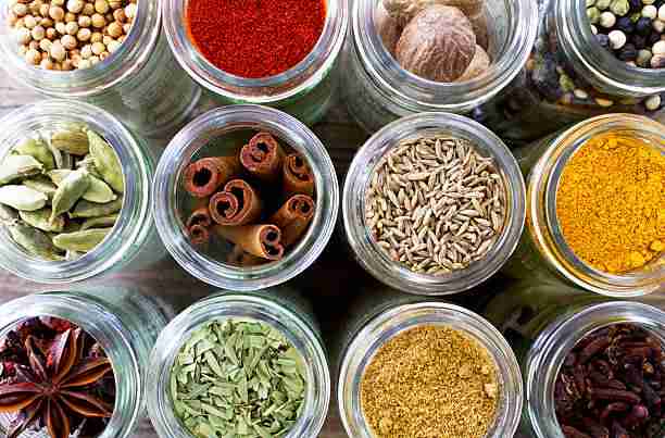 A selection of dried spices and herbs in glass jars shot on a dark wood background. Coriander seed, cloves, cardamom, star anise, thyme, cumin seeds, cinnamon sticks, chilli powder, turmeric, nutmeg and fresh bay leaves. Shot from above.