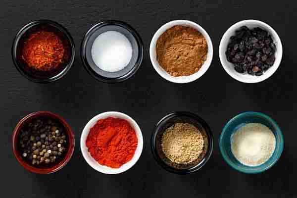 Assorted spices and seasonings in small bowls on dark background, top view.